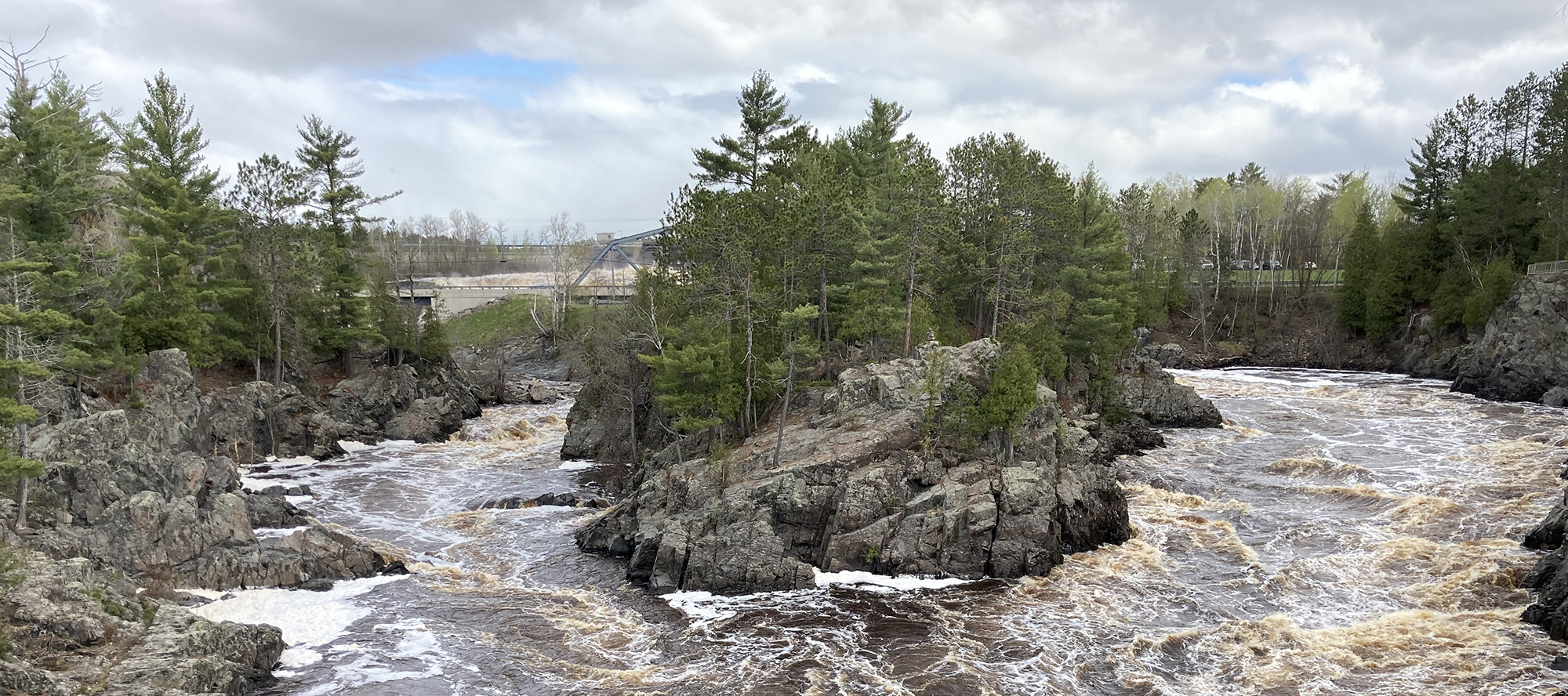 Turbid water flowing over spillway around rocks and trees.