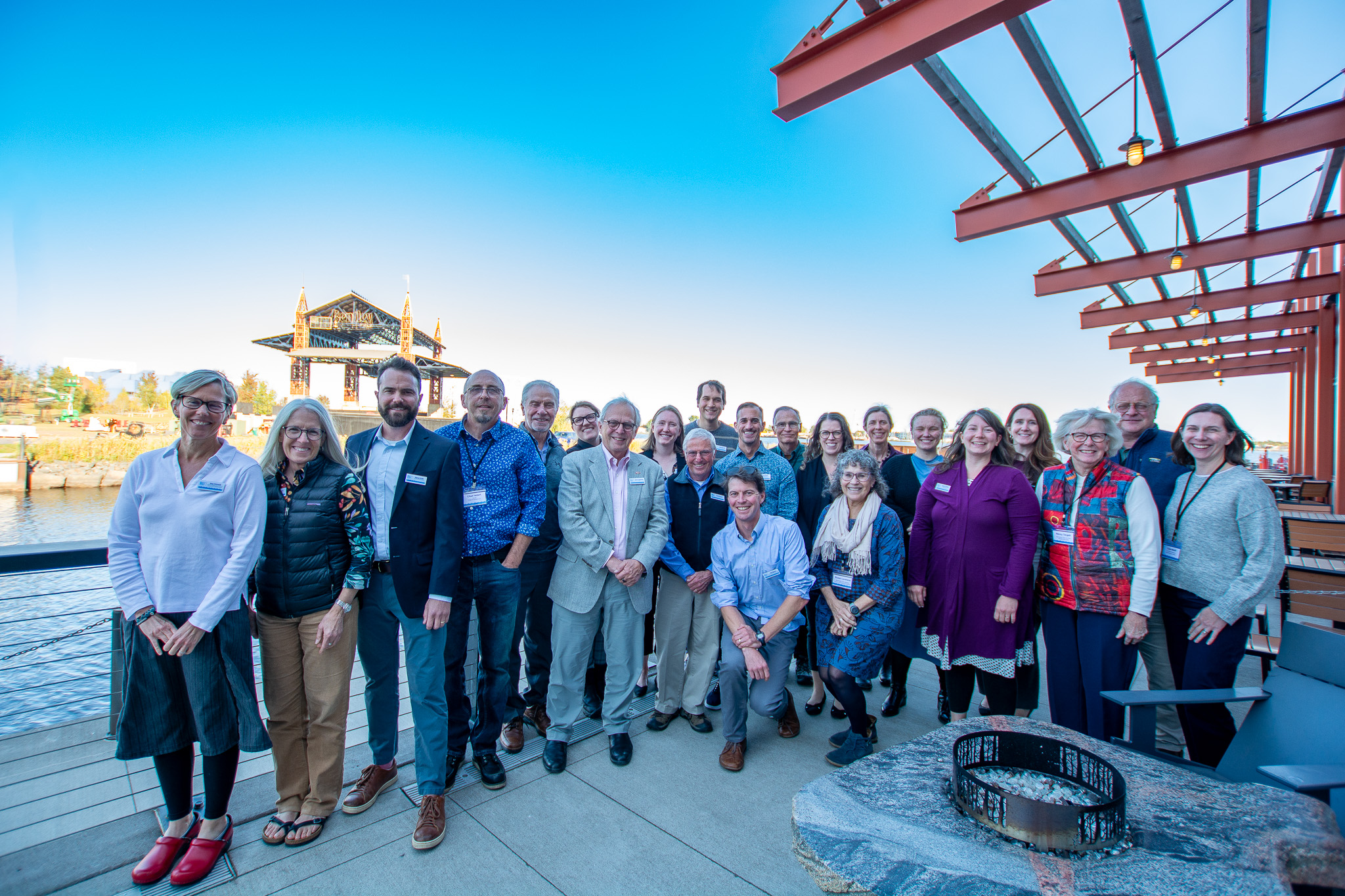 22 people standing shoulder-to-shoulder on a deck near water.