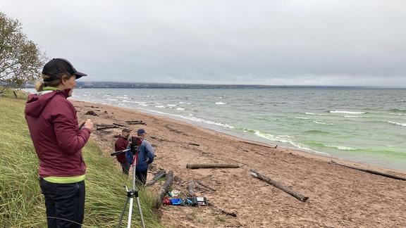 Person standing on dune near camera, two people standing on sandy beach. All three people looking toward Lake Superior and watching the movement of food-grade green dye in the water used to visualize a rip current.