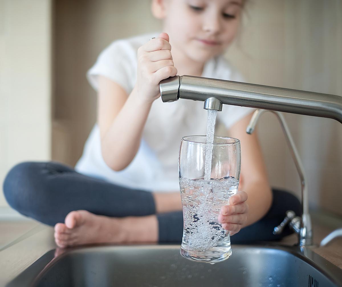 Young child sitting on counter filling glass from water faucet.