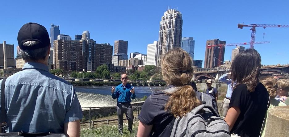 Person talking to a group of people outside with city skyline in the background.