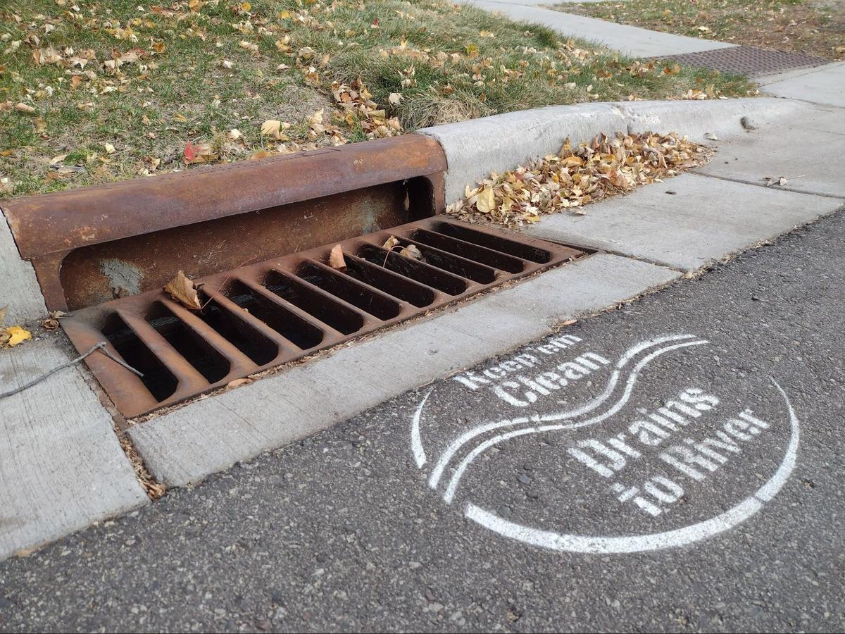 Street curb storm drain without leaves and with stenciled words on pavement that say "Keep them clean. Drains to River."