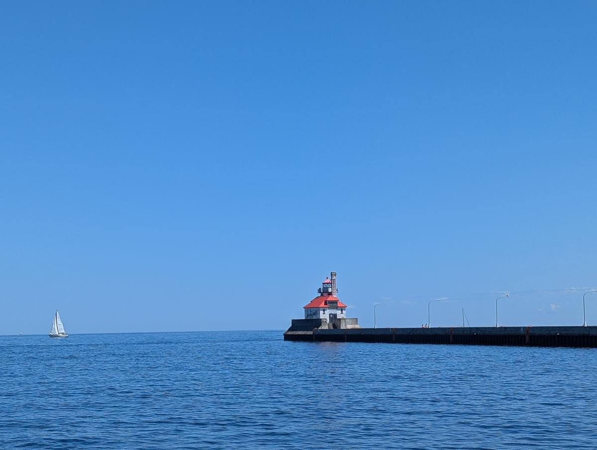 Clear blue skies and calm waters with a sailboat and lighthouse at the entrance of the Duluth port 