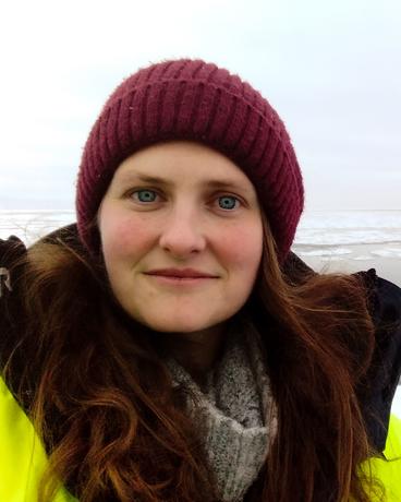 Evelyn Strombom smiles at the camera in cold weather gear with a coast visible behind.